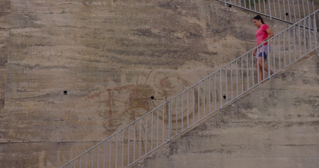 Woman in a pink shirt, shorts, and sneakers descending a large concrete staircase. Useful for depicting urban activities, exercise, daily commute, architecture, and simplicity of urban landscapes. Can be used in campaigns related to fitness, architecture, and lifestyle themes.