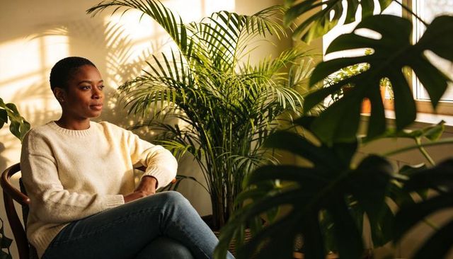 Contemplative woman sitting in sunlit room surrounded by indoor plants and greenery
