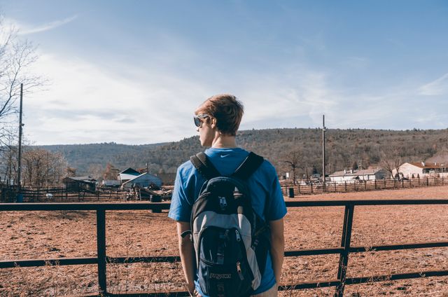 Teenage boy in casual wear standing in a rural setting with a backpack, looking towards distant hills and a picturesque countryside. Ideal for promoting travel, adventure, and youth lifestyle content.