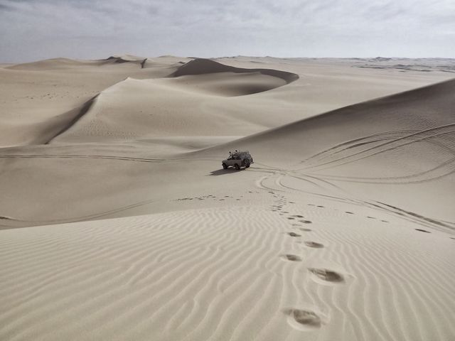 Off-road vehicle driving across expansive desert sand dunes. Footprints trailing from foreground add to sense of adventure and exploration. Ideal for promoting adventure travel, outdoor activities, desert expeditions, and exploring remote, scenic locations.