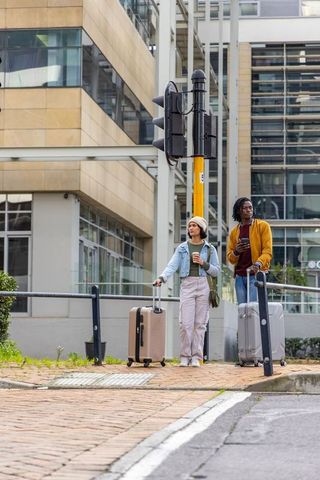 Urban travelers standing with rolling luggage and coffee at city corner near modern offices