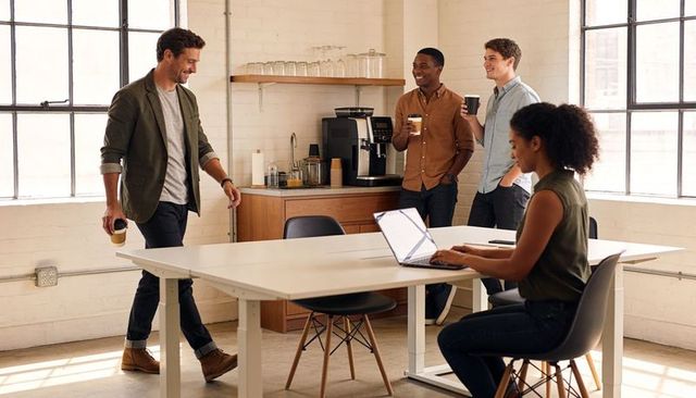 Casual coworking team sharing coffee as woman types on laptop in bright modern workspace