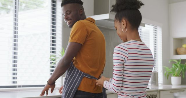 Image of happy african american woman tying apron to her partner. love, relationship, togetherness and spending quality time at home.