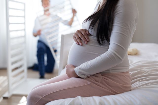 Pregnant woman sitting on bed holding her baby bump while her partner assembles a baby crib in the background. Ideal for use in content related to pregnancy, parenthood, family life, prenatal care, and preparing for a new baby.