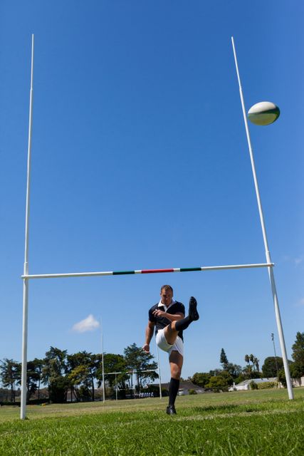Full length of rugby player kicking ball through goal post against clear blue sky