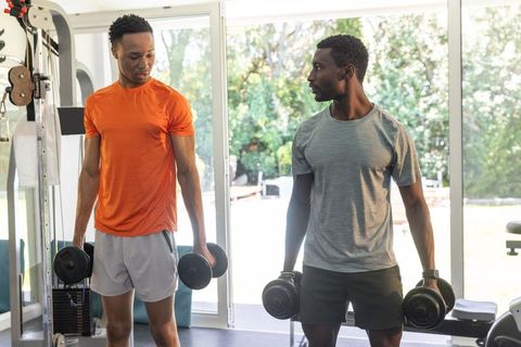 African american men training with dumbbells in sunlit home gym by glass doors