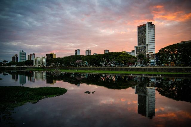 This image shows a city skyline at sunset, with vibrant clouds reflected on the still waters of a nearby river. Tall buildings and lush trees along the water’s edge create a tranquil and picturesque urban scene. Perfect for use in travel brochures, promotional materials for city infrastructure development, or stock photos emphasizing serene urban environments.