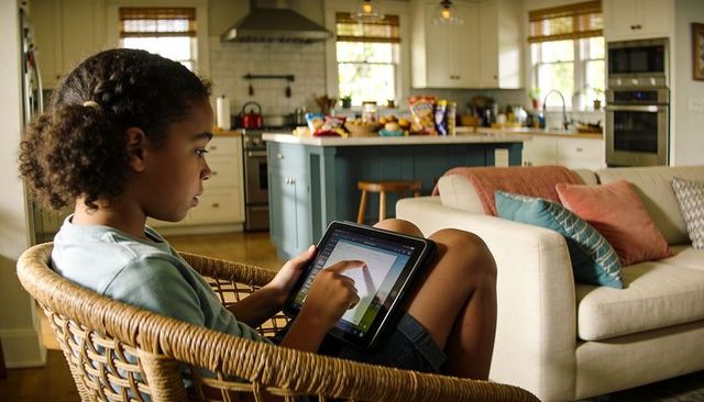 Child using tablet in sunlit open-plan living room, relaxing in wicker chair
