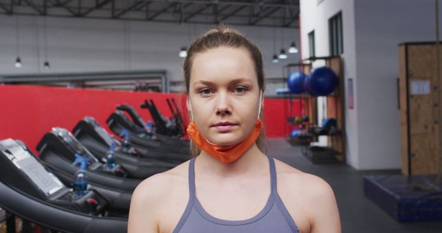 Young woman standing in the gym wearing a lowered face mask beneath her chin and looking at the camera. Treadmills and exercise equipment visible in background. Suitable for use in articles about fitness amidst the pandemic, health precautions, gym culture, or advertisements for gym memberships and wellness programs.