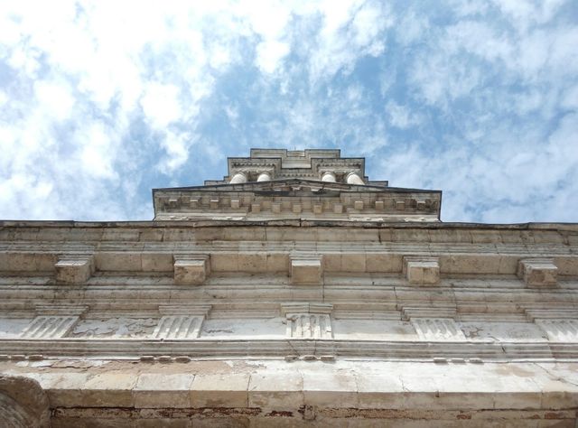 Capturing an upward view of a historical stone building with intricate architectural details set against a clear, cloud-dotted sky. Ideal for use in projects related to travel, history, architecture, and cultural heritage appreciation.