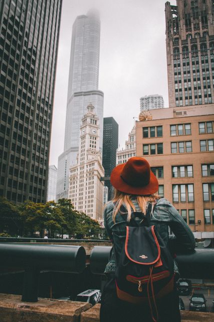 Image shows a woman with a red hat and backpack looking at tall buildings and skyscrapers in an urban cityscape. Great for use in travel blogs, tourism brochures, urban lifestyle themes, and social media content about exploring new cities, solo travel adventures, and city architecture.