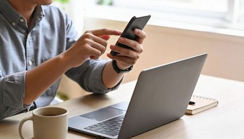 Man using smartphone at sunlit desk with laptop and coffee for remote work
