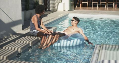 Two friends lounging on inflatable pool float chatting and relaxing on sunny deck