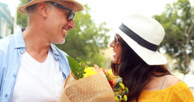 Middle-aged couple standing outdoors, wearing sunglasses and sun hats, smiling at each other. Holding a vibrant floral bouquet, they exude happiness and warmth, suggesting a sunny summer day. Ideal for use in contexts promoting love, relationships, summer activities, outdoor enjoyment, and special celebrations.