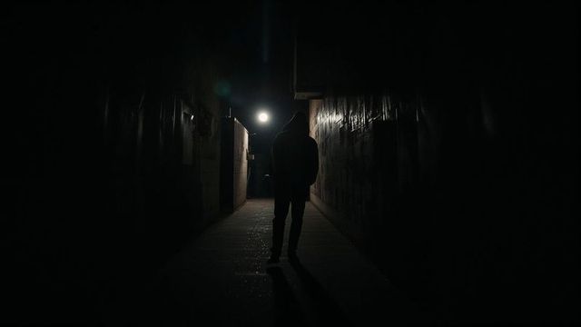 Hooded Figure Walking Through Dark Alley at Night with Sparse Streetlight