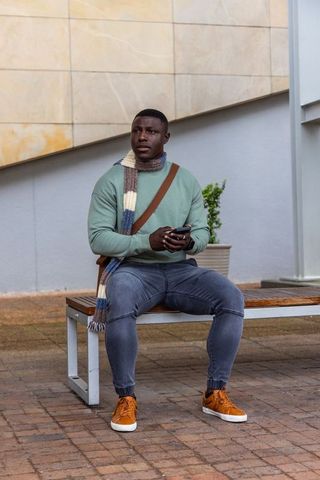 African american man sitting on bench holding smartphone wearing striped scarf urban plaza