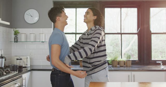 This image shows a young couple embracing and laughing in a modern kitchen with large windows. Can be used for lifestyle blogs, relationship counseling articles, family-oriented promotions, or marketing materials focusing on home and domestic life.