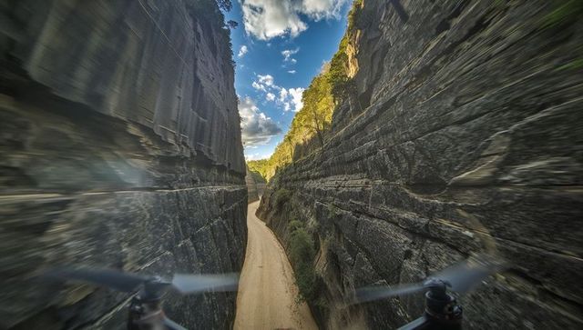 Low-flying drone through narrow stratified canyon over winding dirt track with rotor blur