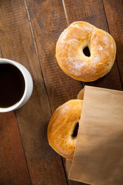 Two fresh bagels and a cup of black coffee on a rustic wooden table. One bagel is partially inside a brown paper bag. Ideal for use in food blogs, breakfast menus, bakery advertisements, and morning routine visuals.