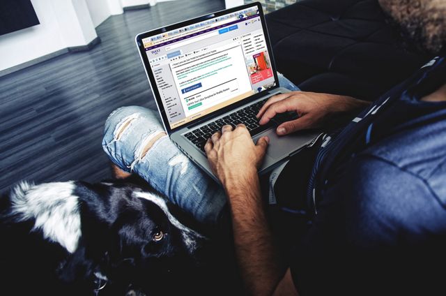 Man sitting and using laptop with a dog by his side inside a modern home. Great for illustrating concepts related to remote work, home office, multitasking, casual working environment, and living with pets.