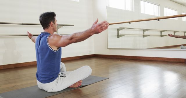 Man practicing yoga in a bright studio with a large mirror, seated on a yoga mat, spreading his arms. Ideal for promoting fitness and wellness programs, yoga studios, meditation retreats, and health blogs.
