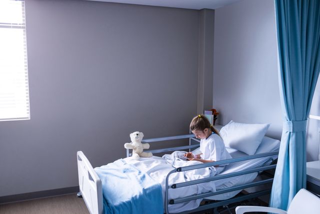 Child patient sitting on hospital bed using digital tablet. Teddy bear placed beside the child. Scene depicts healthcare, recovery, and use of technology in medical settings. Useful for illustrating pediatric care, hospital environments, and the integration of technology in healthcare.