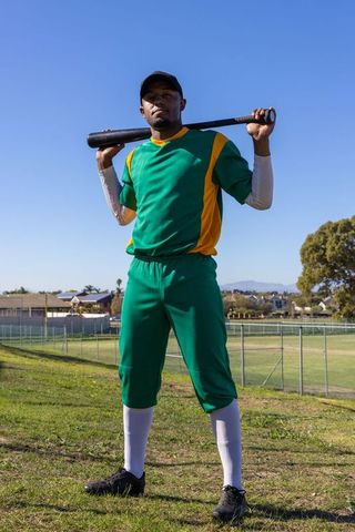African american baseball player standing with bat on suburban field in green uniform