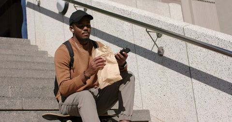 African American man sitting on skateboard on urban steps eating lunch checking smartphone