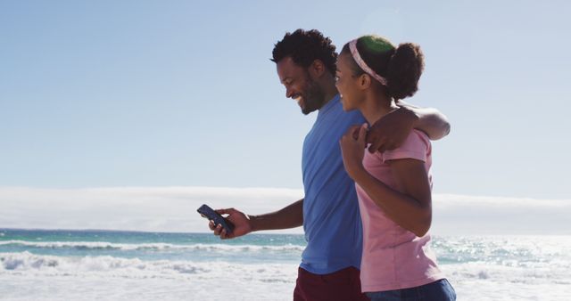 Happy couple enjoying a walk on the beach on a sunny day. They are both dressed in casual attire, and one person is looking at a smartphone while they walk. Ideal for use in projects about relationships, leisure activities, technology, vacations, and outdoor enjoyment.
