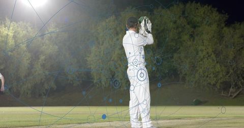 Night cricket wicketkeeper standing under floodlights holding gloves with blue data overlay