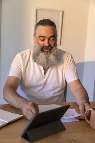 Senior bearded man working at home desk reviewing tablet and paperwork in warm light