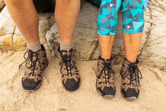 This image depicts the lower legs and feet of a senior couple wearing hiking shoes, sitting on rocks at a sandy beach. Ideal for use in articles or advertisements related to outdoor activities, hiking, adventure travel, senior fitness, and healthy living during retirement.