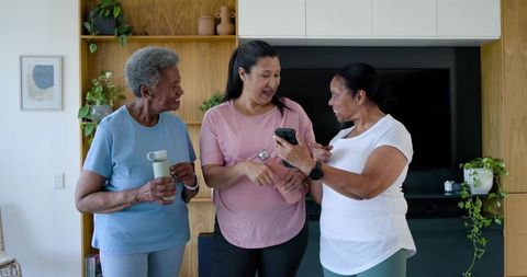 Multigenerational diverse women sharing smartphone and water bottles in cozy living room
