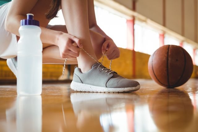 This image shows a woman tying her shoelace while crouching on a basketball court. A water bottle and basketball are visible, indicating a sports or fitness setting. This image can be used for promoting athletic wear, fitness routines, sports events, or health and wellness campaigns.