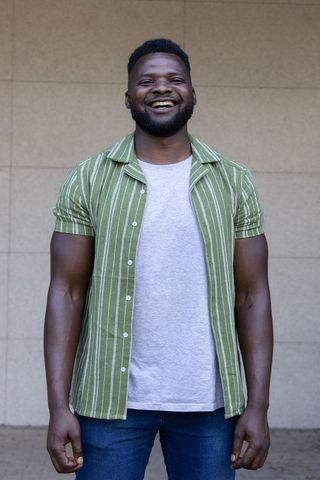 Confident smiling man wearing green striped shirt and jeans standing against neutral wall
