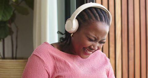 Joyful african american woman wearing white headphones laughing near window cozy interior