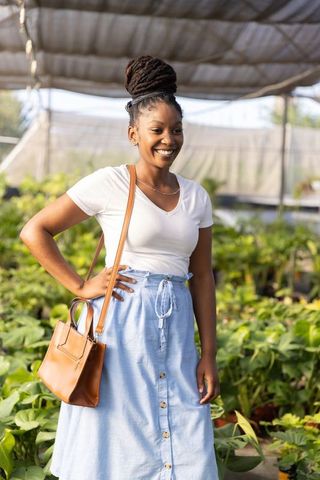 African american woman smiling in greenhouse nursery wearing blue skirt and tan handbag