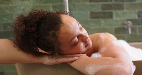 Reclining woman resting on tub rim, soaking in bubble bath with green tile background