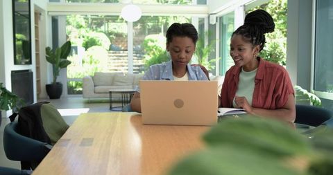 Mother guiding teenage daughter on laptop at bright glass-walled home study table