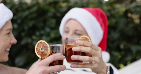 Friends clinking glasses of mulled wine with dried orange garnish wearing santa hats