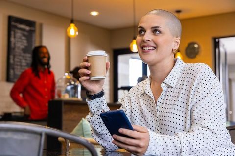 Smiling woman with shaved head holding coffee and phone in bright modern cafe interior