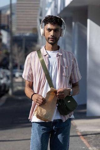 Asian man walking sidewalk wearing headphones carrying paper bag and green crossbody bag