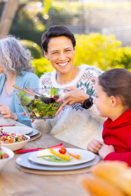 Vertical of happy caucasian mother serving salad in garden with daughter and grandmother, copy space. Family, togetherness, domestic life and happiness concept.