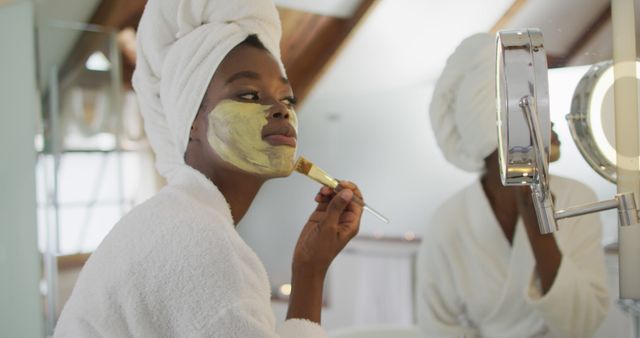 Depicts woman applying face mask while wearing white bathrobe in spa-like bathroom. Suitable for content about skincare routines, beauty treatments, spa experiences, and self-care practices. Can be used in blogs, websites, advertisements, and social media posts promoting wellness and beauty products.