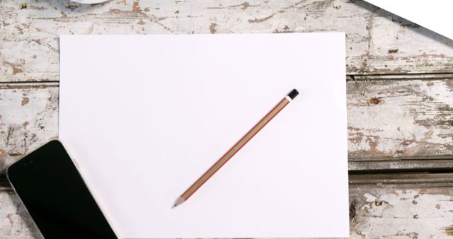 Image shows a top-view of a blank white paper with a pencil placed on it, alongside a smartphone, all situated on a rustic wooden desk. Ideal for conveying creativity, brainstorming, minimalism, simplicity, workspace organization, remote working themes or art and craft projects.