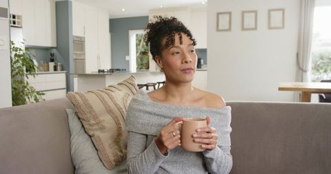 Relaxing African American woman holding mug on sofa in modern minimalist open-plan home
