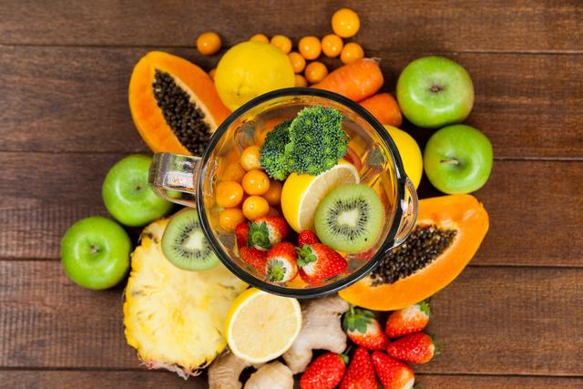 Overhead view of a variety of fresh fruits and vegetables in a blender on a wooden table. This vibrant and colorful arrangement includes kiwi, strawberries, papaya, apples, lemon, pineapple, ginger, carrots, and broccoli. Ideal for promoting healthy eating, diet plans, smoothie recipes, and nutrition blogs.