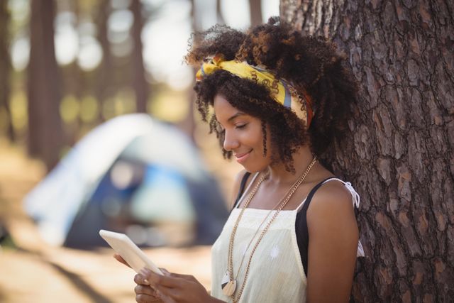 Young woman standing by tree trunk at campsite, using smartphone. Ideal for themes related to outdoor activities, camping, technology in nature, leisure time, and modern lifestyle.