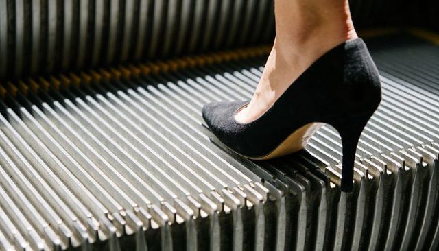 Stiletto stepping on grooved escalator tread close-up, black suede pump ankle detail