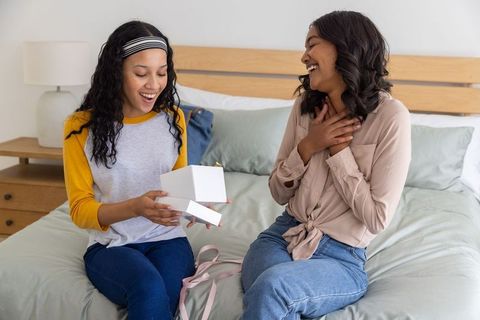 Mother and teenage daughter opening gift on bed, laughing and sharing joyful moment